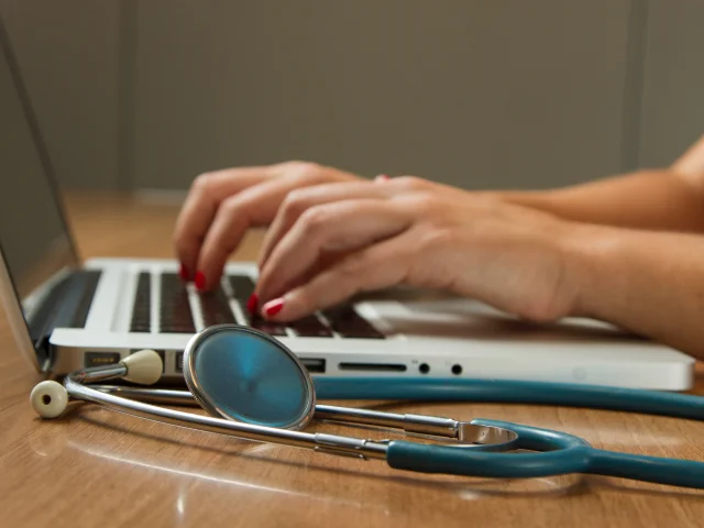 Mains d'un professionnel de santé tapant sur le clavier d'un ordinateur portable avec un stéthoscope.