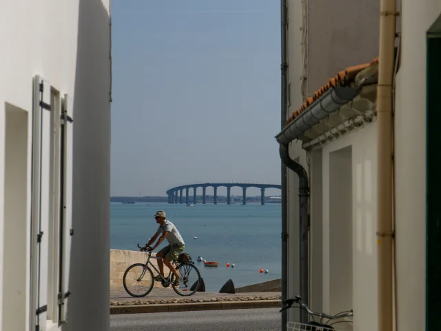 Personne faisant du vélo et visible entre deux murs d'une venelle de Rivedoux avec la mer et le pont de l'île de Ré en arrière plan