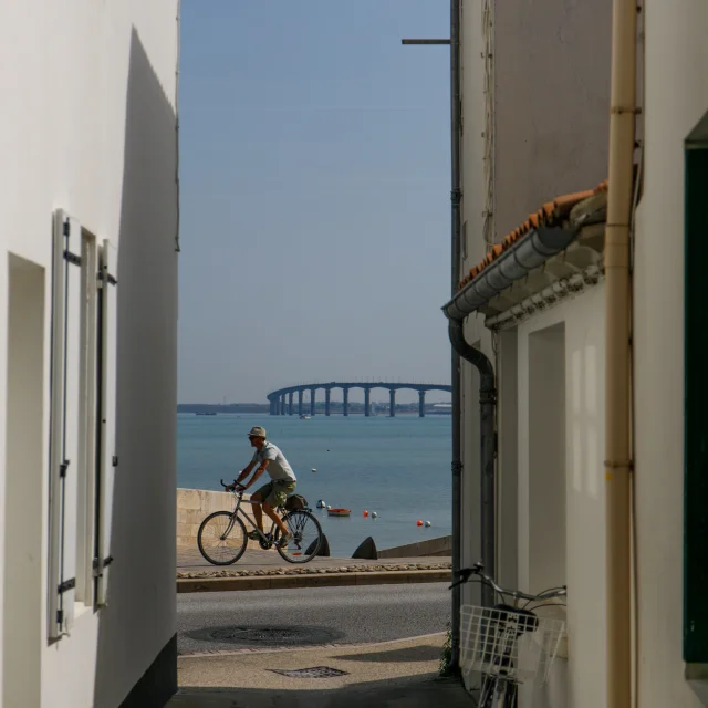 Personne faisant du vélo et visible entre deux murs d'une venelle de Rivedoux avec la mer et le pont de l'île de Ré en arrière plan