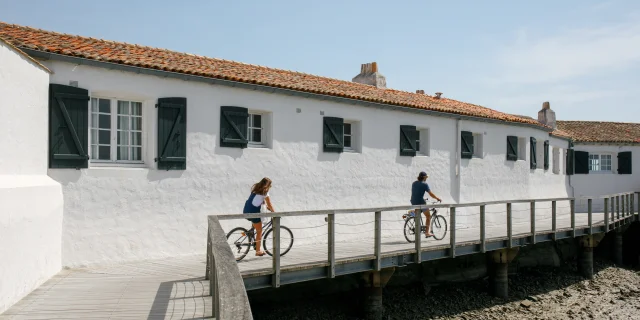 Deux personnes à vélo sur une passerelle en bois près d'un bâtiment blanc.