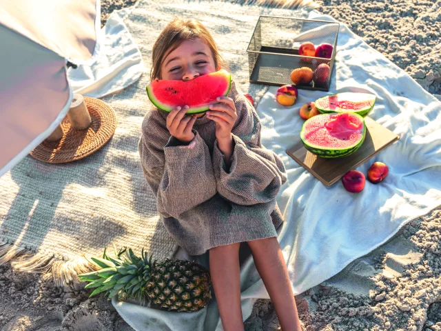 Petite fille assise sur une couverture de plage mangeant une pastèque.