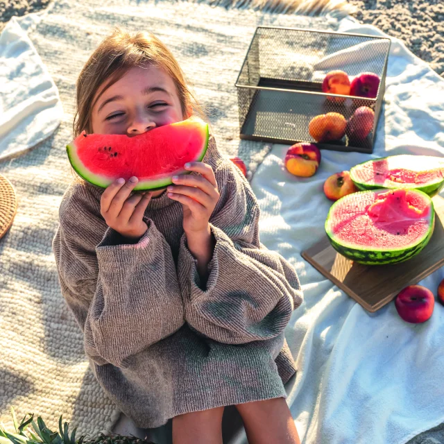 Petite fille assise sur une couverture de plage mangeant une pastèque.