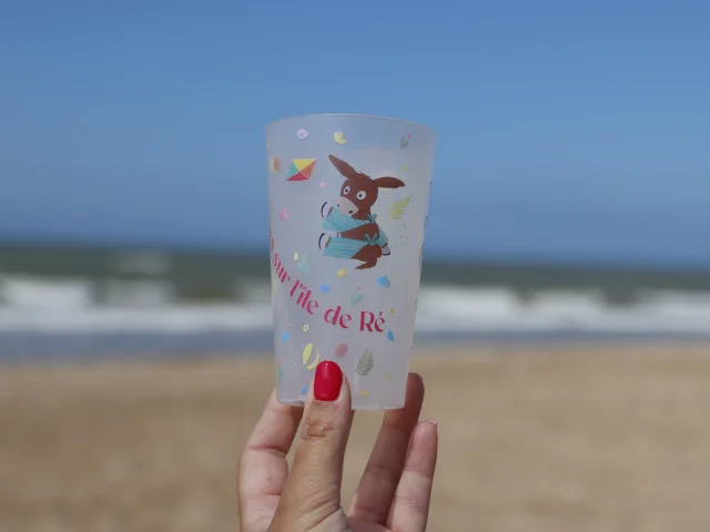 Gobelet transparent avec dessin d'un âne, tenu devant la plage.