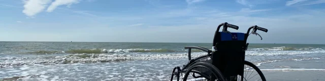 Un fauteuil roulant est placé sur le sable, près du bord de mer, sous un ciel bleu.