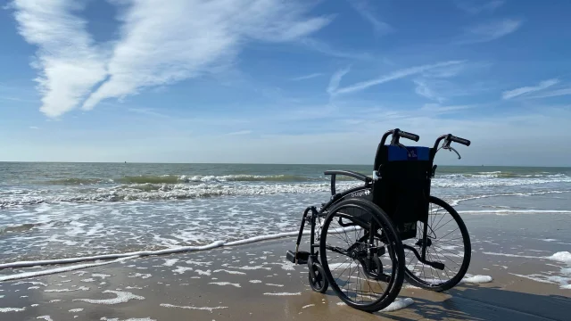 Un fauteuil roulant est placé sur le sable, près du bord de mer, sous un ciel bleu.