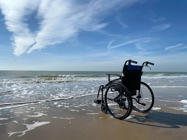 Un fauteuil roulant est placé sur le sable, près du bord de mer, sous un ciel bleu.