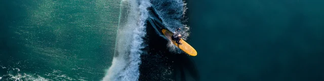 Surfeur sur une grande vague vue aérienne, la planche jaune contraste avec l'eau.