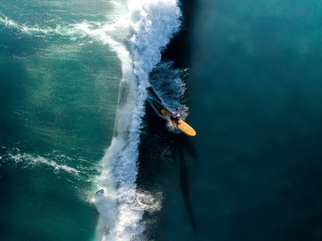 Surfeur sur une grande vague vue aérienne, la planche jaune contraste avec l'eau.