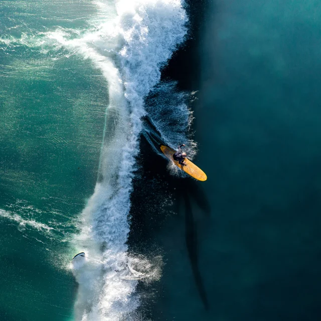 Surfeur sur une grande vague vue aérienne, la planche jaune contraste avec l'eau.